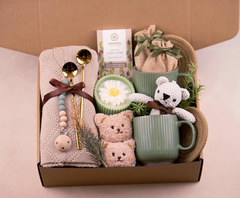 May include: A gift box containing baby-related items. It features a beige blanket, gold spoons, a dummy, two green mugs, a white teddy bear, two small teddy bear faces, a candle, and coffee. The box is brown.