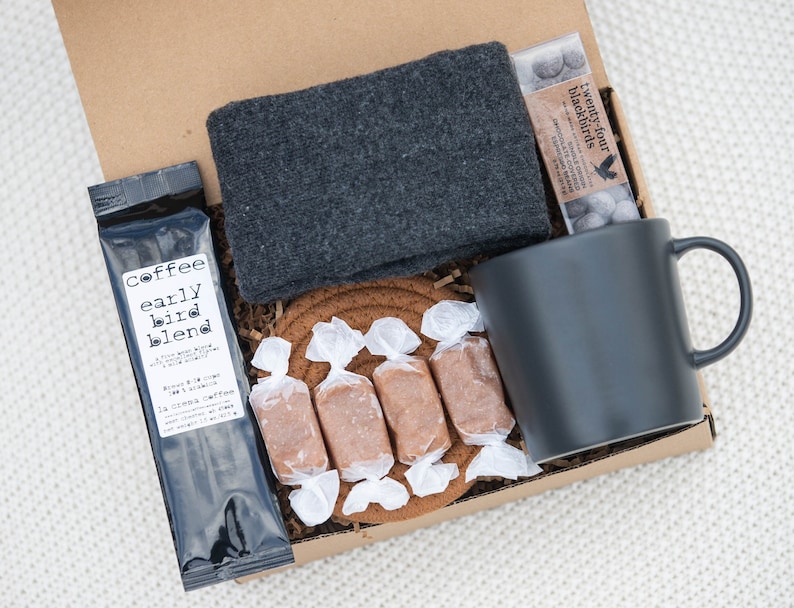 May include: A gift box filled with a black mug, a bag of coffee labelled "Coffee Early Bird Blend", a pair of grey socks, a bag of chocolate candies labelled "Twenty-Four Blackbirds", and caramel candies wrapped in white paper.
