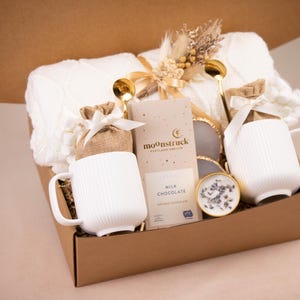 May include: A gift box filled with a white knitted blanket, two white ribbed mugs, gold spoons, a chocolate bar, and a small candle. The box is brown and tied with a ribbon, creating a cozy and inviting aesthetic.