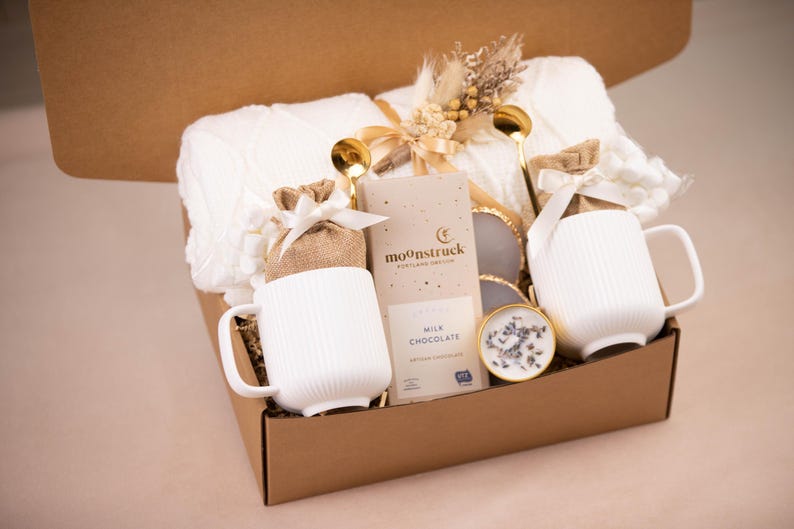 May include: A gift box filled with a white knitted blanket, two white ribbed mugs, gold spoons, a chocolate bar, and a small candle. The box is brown and tied with a ribbon, creating a cozy and inviting aesthetic.