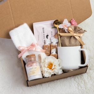 May include: A gift box filled with self-care items. It includes a white mug, a white towel with a pink bow, a bottle of bath salts, a gold spoon, a face mask, candies, and a small burlap bag with a bow, all arranged in a brown cardboard box.