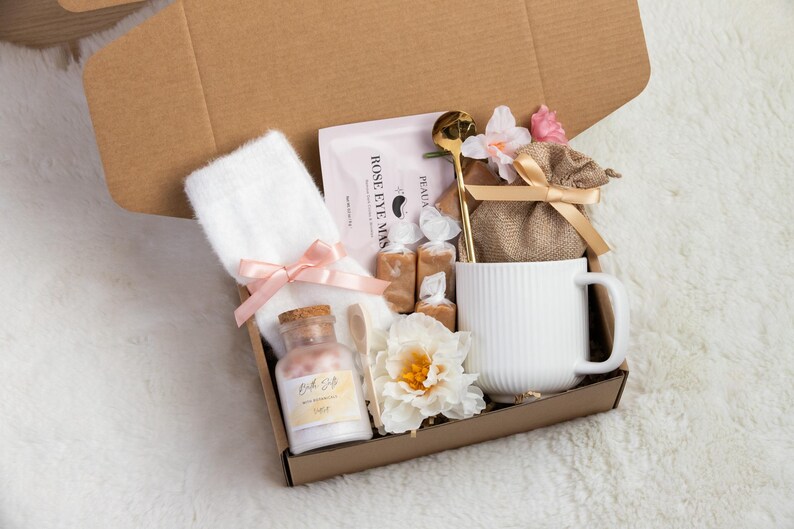 May include: A gift box filled with self-care items. It includes a white mug, a white towel with a pink bow, a bottle of bath salts, a gold spoon, a face mask, candies, and a small burlap bag with a bow, all arranged in a brown cardboard box.