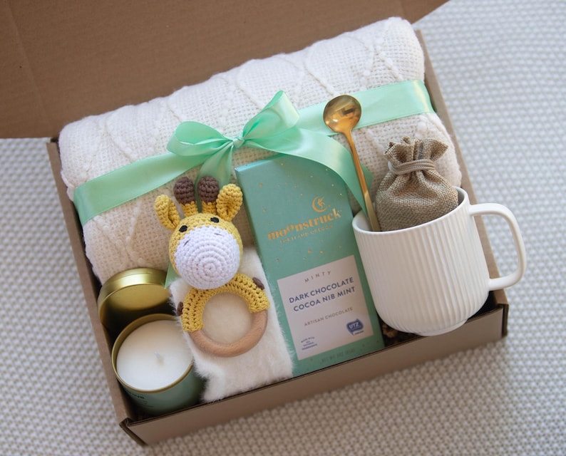 Op de afbeelding: Een cadeaudoos gevuld met artikelen voor een nieuwe ouder. De doos bevat een wit gebreid dekentje, een geel gehaakt giraffenknuffeltje, een kaars, een mok, een zakje cacao-mix en een gouden lepel.