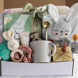 May include: A gift box filled with self-care items and baby products. Includes a white mug, a bunny-themed soft book, a crochet bunny, face masks, a wooden teether, a scrunchie, a gold spoon, and a bar of soap. The box is tied with a green ribbon.