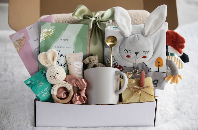 May include: A gift box filled with self-care items and baby products. Includes a white mug, a bunny-themed soft book, a crochet bunny, face masks, a wooden teether, a scrunchie, a gold spoon, and a bar of soap. The box is tied with a green ribbon.