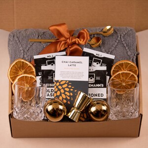 May include: A gift box containing a grey cable-knit blanket tied with an orange ribbon, two clear textured glass tumblers, two gold-toned spherical ice molds, a gold jigger, two gold spoons, dried orange slices, and Chai Caramel Latte drink mix.