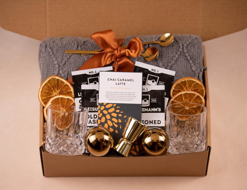 May include: A gift box containing a grey cable-knit blanket tied with an orange ribbon, two clear textured glass tumblers, two gold-toned spherical ice molds, a gold jigger, two gold spoons, dried orange slices, and Chai Caramel Latte drink mix.