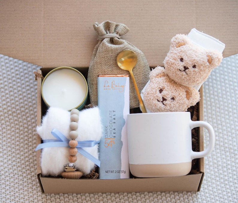 Op de afbeelding: Een cadeaudoos gevuld met artikelen voor een nieuwe ouder. De doos bevat een witte mok, een kaars, een reep donkere chocolade met zeezout, een houten bijtring, een witte babysok met een bruin beren gezicht, een klein tasje en een gouden lepel.
