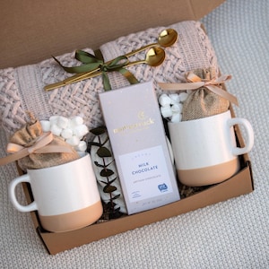 May include: A gift box filled with a cream-colored knit blanket, two white mugs with tan bottoms, a bar of milk chocolate, two small burlap bags, marshmallows, and two gold spoons. The box is tied with a green ribbon.