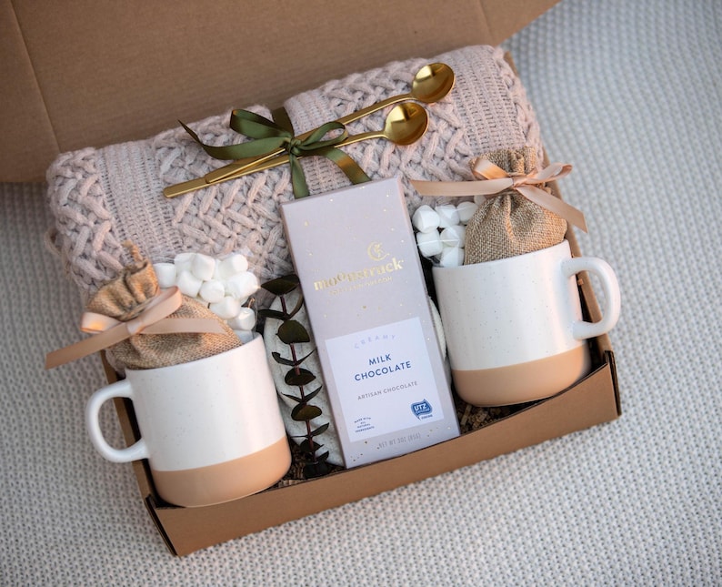 May include: A gift box filled with a cream-colored knit blanket, two white mugs with tan bottoms, a bar of milk chocolate, two small burlap bags, marshmallows, and two gold spoons. The box is tied with a green ribbon.