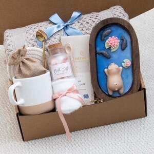May include: A gift box filled with self-care items. Includes a white mug with a tan base, a jar of bath salts, a small bag, a gold spoon, a bar of chocolate, a blue bow, a small white towel, and a wooden decorative piece with a pig figurine.