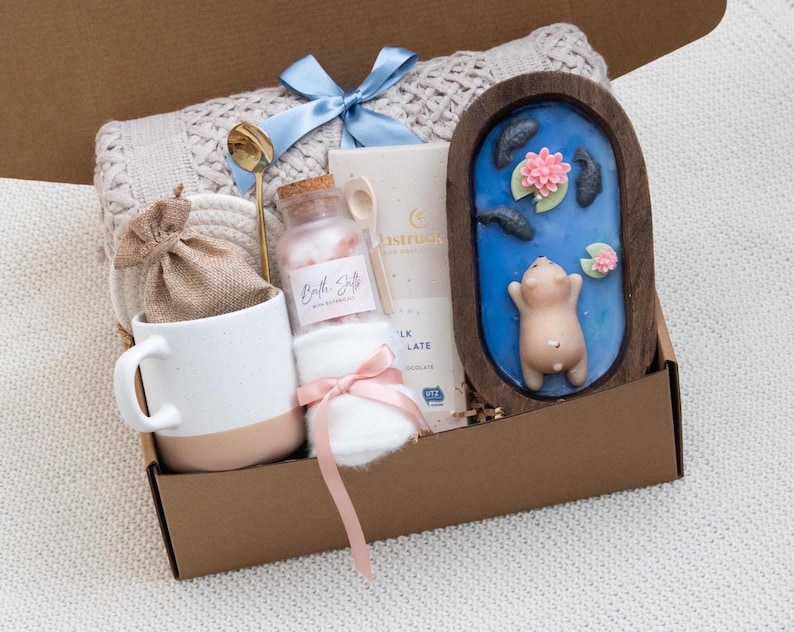 May include: A gift box filled with self-care items. Includes a white mug with a tan base, a jar of bath salts, a small bag, a gold spoon, a bar of chocolate, a blue bow, a small white towel, and a wooden decorative piece with a pig figurine.