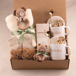 May include: A gift box filled with baby items and treats. Includes a teddy bear rattle, blanket, socks, two teddy bear booties, a chocolate bar, two mugs that say "daddy" and "mommy", two gold spoons, and two small burlap bags.