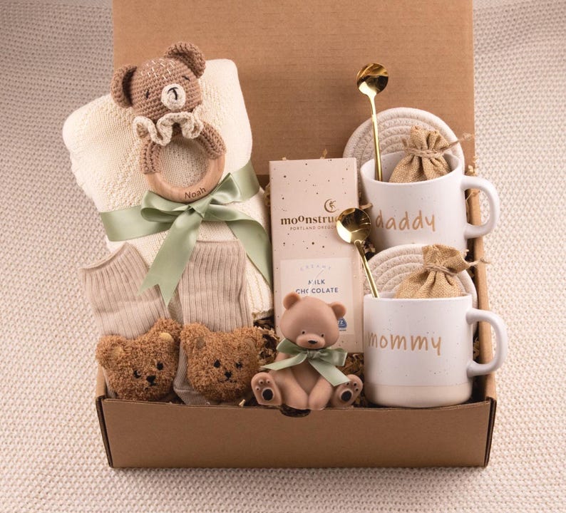 May include: A gift box filled with baby items and treats. Includes a teddy bear rattle, blanket, socks, two teddy bear booties, a chocolate bar, two mugs that say "daddy" and "mommy", two gold spoons, and two small burlap bags.