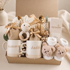 May include: A gift box filled with baby items and gifts for parents. Includes two white mugs with "mommy" and "daddy" printed on them, a crochet bunny rattle, teddy bear socks, and a chocolate bar.