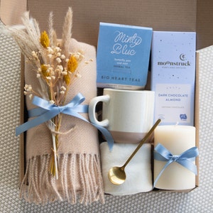 Peut inclure: Une bo&icirc;te-cadeau remplie d'une couverture douillette, une tasse, une bougie, une bo&icirc;te de th&eacute; aux herbes et une bo&icirc;te de chocolat noir aux amandes artisanal. La bo&icirc;te est d&eacute;cor&eacute;e d'un ruban bleu et d'un bouquet de fleurs s&eacute;ch&eacute;es.