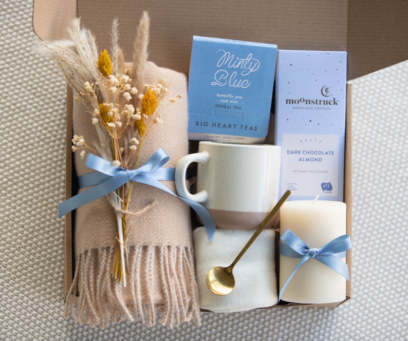 Peut inclure: Une bo&icirc;te-cadeau remplie d'une couverture douillette, une tasse, une bougie, une bo&icirc;te de th&eacute; aux herbes et une bo&icirc;te de chocolat noir aux amandes artisanal. La bo&icirc;te est d&eacute;cor&eacute;e d'un ruban bleu et d'un bouquet de fleurs s&eacute;ch&eacute;es.