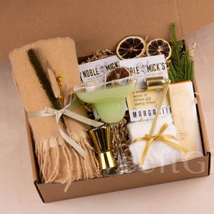 May include: A gift box filled with margarita cocktail essentials. Includes a margarita glass with a green drink, a gold jigger, a gold bar tool, a beige scarf, and other items. The box is brown and contains dried citrus slices.