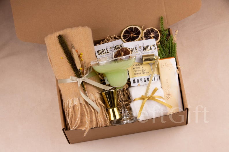 May include: A gift box filled with margarita cocktail essentials. Includes a margarita glass with a green drink, a gold jigger, a gold bar tool, a beige scarf, and other items. The box is brown and contains dried citrus slices.