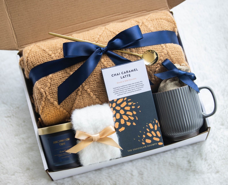 K&ouml;nnte beinhalten: Eine Geschenkbox mit einer beigefarbenen Decke, marineblauem Band, goldenem L&ouml;ffel, einer Tasse, einer Kerze, einer Schokoladentafel und einem kleinen, flauschigen wei&szlig;en Gegenstand. Auf der Schokoladentafel steht "Chai Caramel Latte". Auf der Kerze steht "Birchwood Tide".