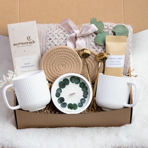 May include: A gift box filled with a white ribbed mug, chocolate bar, candle, coffee, and a knitted blanket. The box also contains two gold spoons and two woven coasters. The candle is decorated with green leaves.