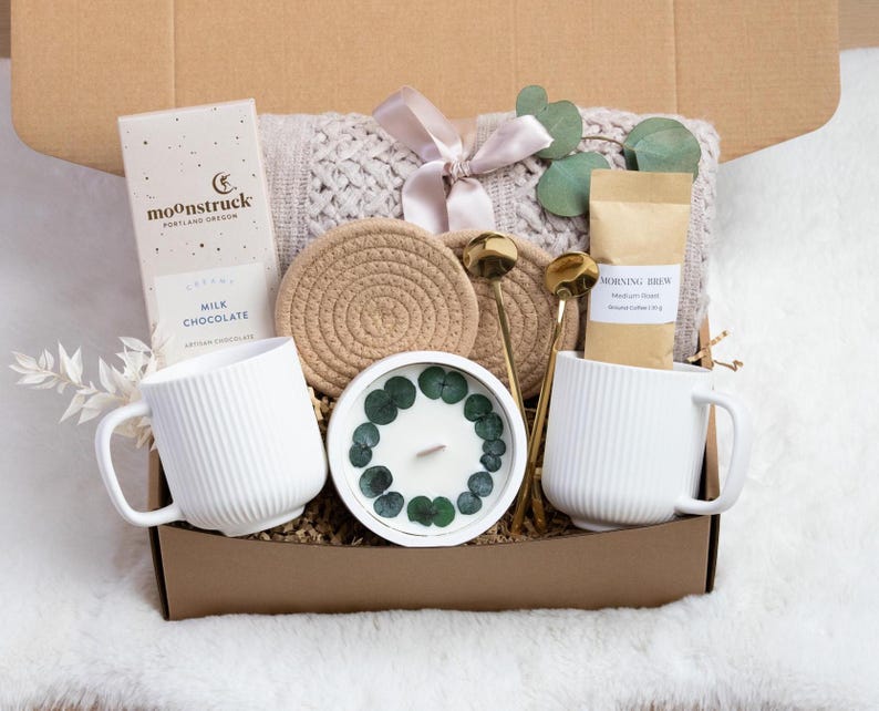 May include: A gift box filled with a white ribbed mug, chocolate bar, candle, coffee, and a knitted blanket. The box also contains two gold spoons and two woven coasters. The candle is decorated with green leaves.
