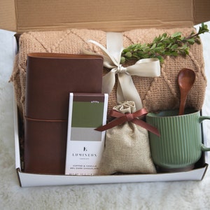 K&ouml;nnte beinhalten: Eine Geschenkbox gef&uuml;llt mit einem braunen Lederjournal, einer gr&uuml;nen Tasse, einem Holzl&ouml;ffel, einer Schokoladentafel, einem kleinen Jutesack und einer beigefarbenen Decke. Die Box ist mit einem cremefarbenen Band gebunden und mit Gr&uuml;nzeug verziert.