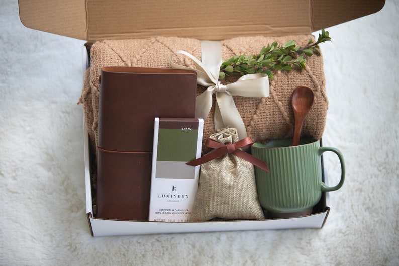 K&ouml;nnte beinhalten: Eine Geschenkbox gef&uuml;llt mit einem braunen Lederjournal, einer gr&uuml;nen Tasse, einem Holzl&ouml;ffel, einer Schokoladentafel, einem kleinen Jutesack und einer beigefarbenen Decke. Die Box ist mit einem cremefarbenen Band gebunden und mit Gr&uuml;nzeug verziert.
