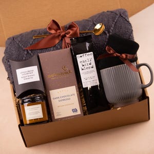 May include: A brown gift box filled with a cosy grey knitted scarf, a mug, a candle, a bag of dark chocolate espresso beans, and a bag of coffee labelled "Coffee Early Bird Blend".