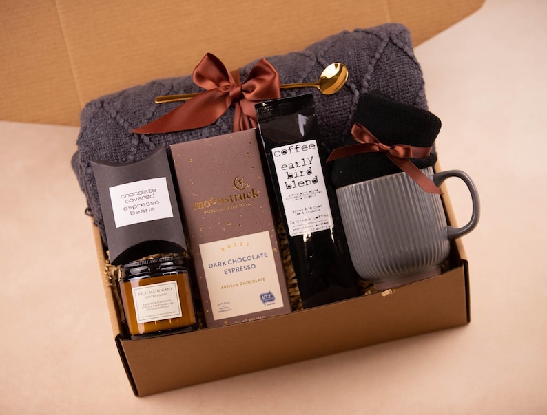 May include: A brown gift box filled with a cosy grey knitted scarf, a mug, a candle, a bag of dark chocolate espresso beans, and a bag of coffee labelled "Coffee Early Bird Blend".