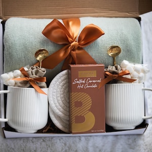 May include: Gift box with a light green blanket, two white ribbed mugs, gold spoons, marshmallows, and a box of salted caramel hot chocolate. The box is tied with an orange ribbon.