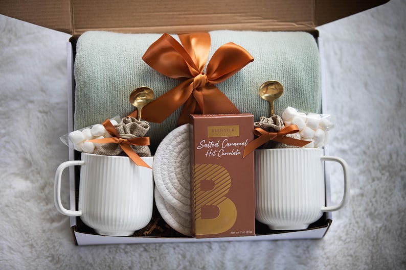 May include: Gift box with a light green blanket, two white ribbed mugs, gold spoons, marshmallows, and a box of salted caramel hot chocolate. The box is tied with an orange ribbon.