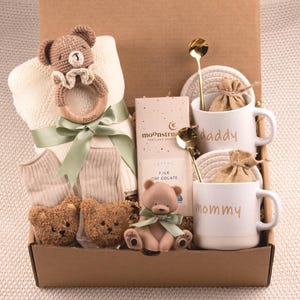 May include: A baby gift basket filled with a crochet teddy bear rattle, a blanket, socks, two plush teddy bears, a small teddy bear figurine, two mugs with the words "daddy" and "mommy", two gold spoons, and chocolate.