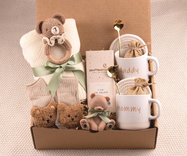 May include: A baby gift basket filled with a crochet teddy bear rattle, a blanket, socks, two plush teddy bears, a small teddy bear figurine, two mugs with the words "daddy" and "mommy", two gold spoons, and chocolate.