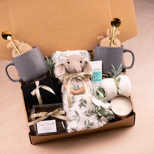 May include: A gift box filled with various items, including two gray ribbed mugs with gold spoons, a black item with a bow, a crocheted elephant, a blanket with a leaf pattern, a tube of hand cream, a candle, and a dark chocolate bar.