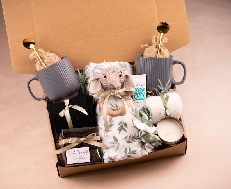 May include: A gift box filled with various items, including two gray ribbed mugs with gold spoons, a black item with a bow, a crocheted elephant, a blanket with a leaf pattern, a tube of hand cream, a candle, and a dark chocolate bar.