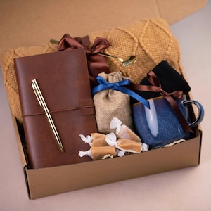 May include: A brown cardboard box filled with a variety of gifts, including a brown leather journal with a gold pen, a blue ceramic mug, a small hessian bag with a brown ribbon, a knitted scarf, and caramel sweets.