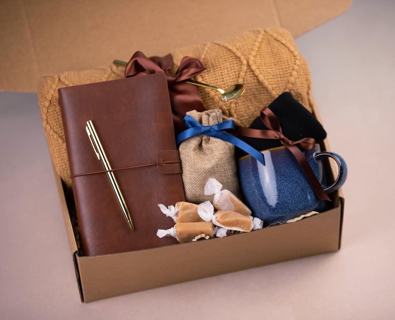 May include: A brown cardboard box filled with a variety of gifts, including a brown leather journal with a gold pen, a blue ceramic mug, a small hessian bag with a brown ribbon, a knitted scarf, and caramel sweets.