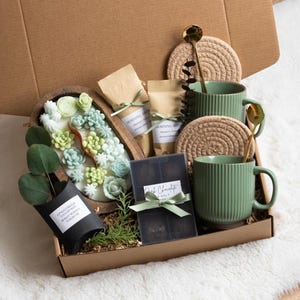 May include: A gift box filled with various items. It includes two green mugs, two gold spoons, two woven coasters, two small bags with text, a wooden tray with succulent-shaped soaps, a black container with text, and a small plant.