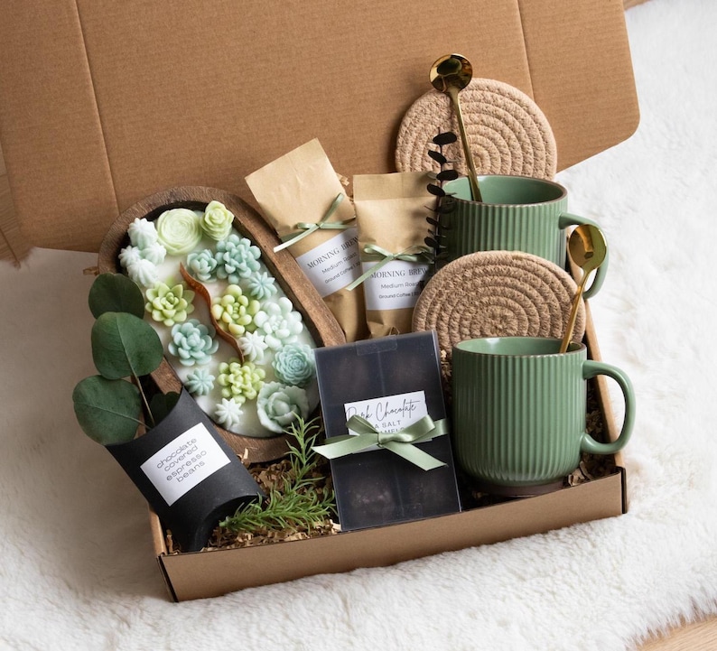 May include: A gift box filled with various items. It includes two green mugs, two gold spoons, two woven coasters, two small bags with text, a wooden tray with succulent-shaped soaps, a black container with text, and a small plant.