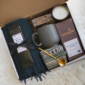 May include: A gift box filled with cosy items for a relaxing evening. It includes a dark green scarf, a grey mug, a small candle in a brown jar, a package of chocolate espresso, a gold spoon, and a package of coffee labelled "early bird blend".