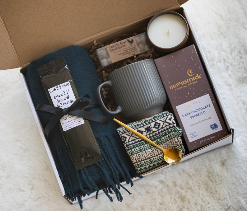 May include: A gift box filled with cosy items for a relaxing evening. It includes a dark green scarf, a grey mug, a small candle in a brown jar, a package of chocolate espresso, a gold spoon, and a package of coffee labelled "early bird blend".
