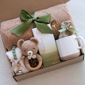 Op de afbeelding: Een cadeaudoos gevuld met een bruin gebreid dekentje, een gehaakte teddybeer met een houten rammelaar, een groene kralenketting, een reep pure chocolade, een mok, een klein zakje van bruin materiaal en een gouden lepel.