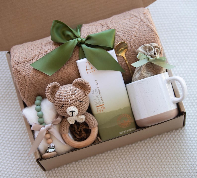 Op de afbeelding: Een cadeaudoos gevuld met een bruin gebreid dekentje, een gehaakte teddybeer met een houten rammelaar, een groene kralenketting, een reep pure chocolade, een mok, een klein zakje van bruin materiaal en een gouden lepel.