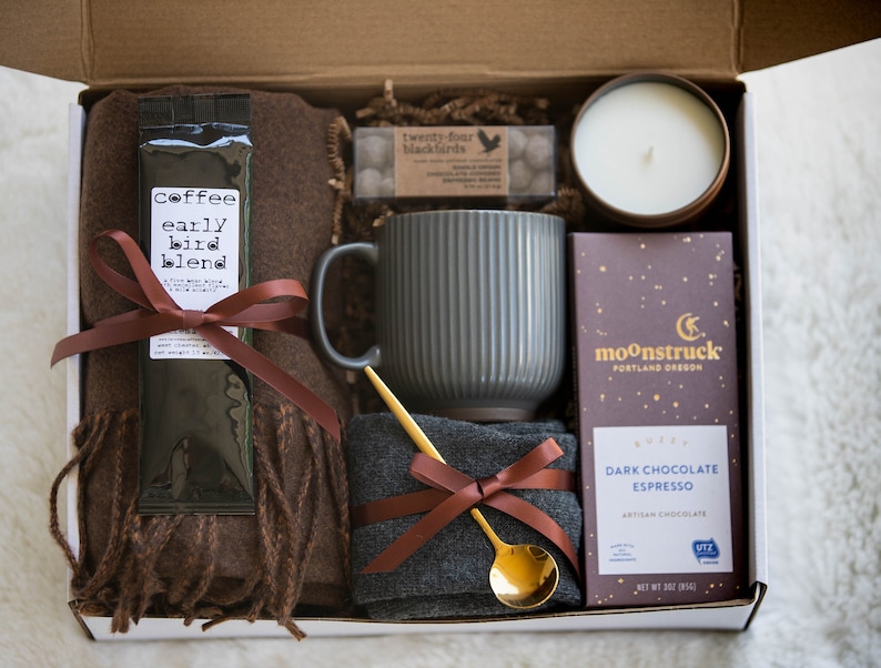 May include: A gift box filled with a brown scarf, a gray mug, a gold spoon, a package of coffee labeled 'coffee early bird blend', a candle, and a bar of dark chocolate espresso. The chocolate bar is labeled 'moonstruck' and 'dark chocolate espresso'. The box is lined with white paper.