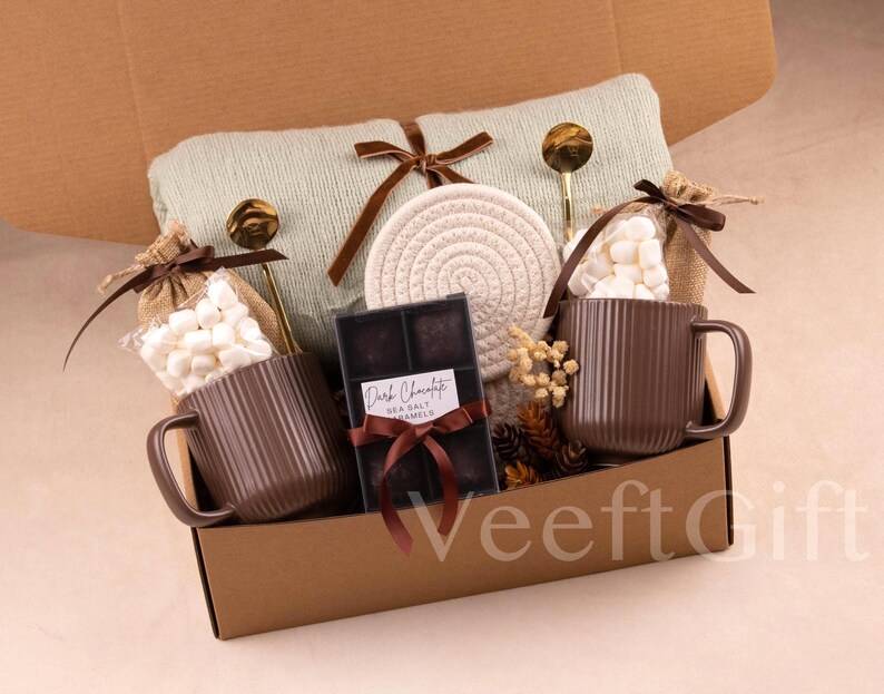 Puede incluir: Una caja de regalo con tem&aacute;tica acogedora. Incluye dos tazas marrones acanaladas, malvaviscos, chocolate negro, una manta verde claro y un posavasos tejido. Cucharas doradas y detalles decorativos completan el conjunto.