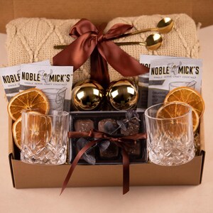May include: A gift box containing a beige knit blanket tied with a brown ribbon, two gold spoons, two Noble Mick's cocktail mix packets, two gold spheres, two dried orange slices, two crystal-cut glass tumblers, and a box of chocolates with a ribbon.