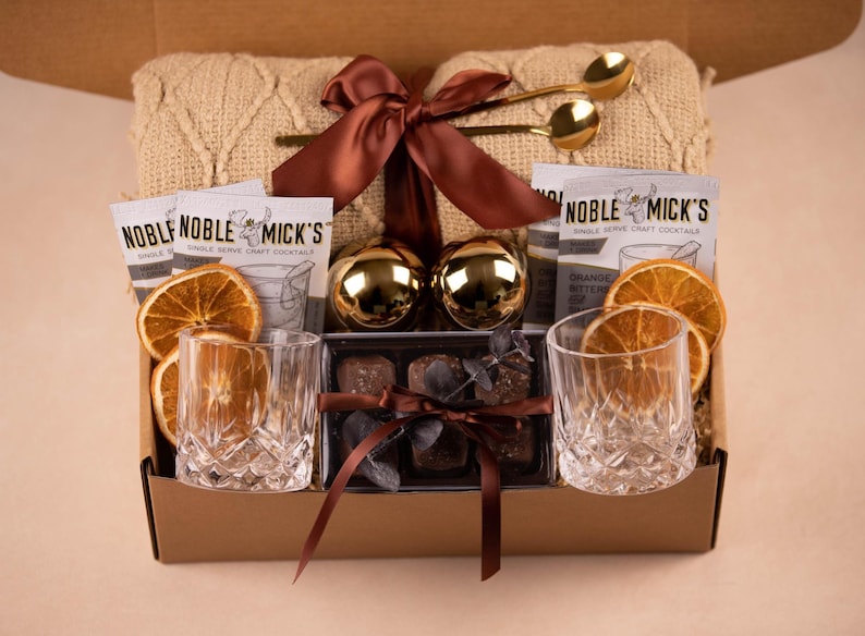 May include: A gift box containing a beige knit blanket tied with a brown ribbon, two gold spoons, two Noble Mick's cocktail mix packets, two gold spheres, two dried orange slices, two crystal-cut glass tumblers, and a box of chocolates with a ribbon.