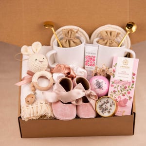 May include: A gift box containing baby and self-care items. It features a crochet bunny, baby shoes, a wooden teether, chocolate, a bath bomb, a candle, two mugs, and hand cream. The box is brown.