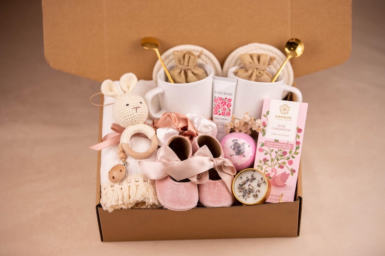 May include: A gift box containing baby and self-care items. It features a crochet bunny, baby shoes, a wooden teether, chocolate, a bath bomb, a candle, two mugs, and hand cream. The box is brown.